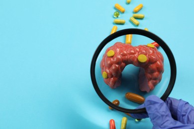 Photo of Endocrinologist looking at model of thyroid gland and capsules through magnifying glass on light blue background, closeup. Space for text