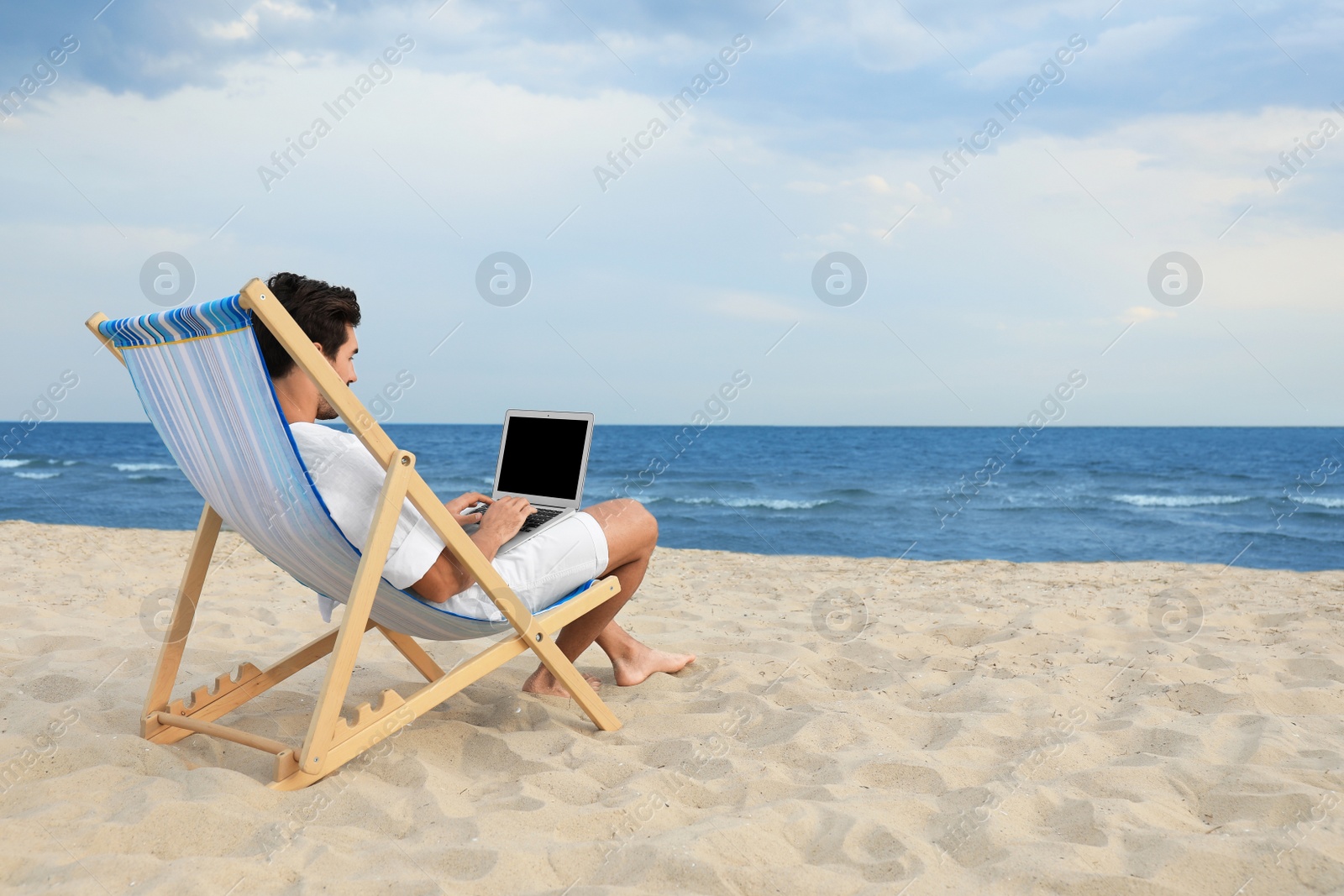 Photo of Young man with laptop sitting in deck chair on sea beach. Space for text