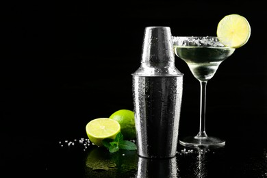 Photo of Metal shaker, delicious cocktail, limes and mint on black mirror surface, space for text