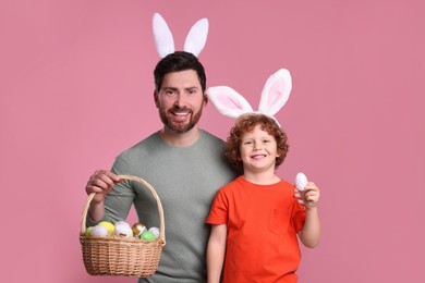 Happy father and son in cute bunny ears headbands holding Easter eggs on pink background