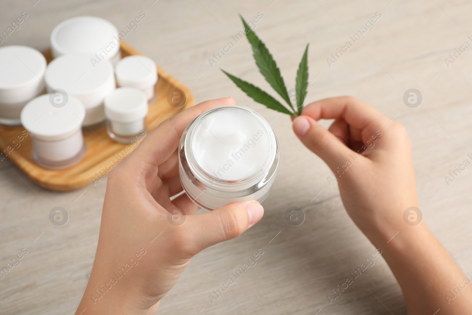 Photo of Woman holding jar of hemp cream and green leaf at wooden table, closeup. Natural cosmetics