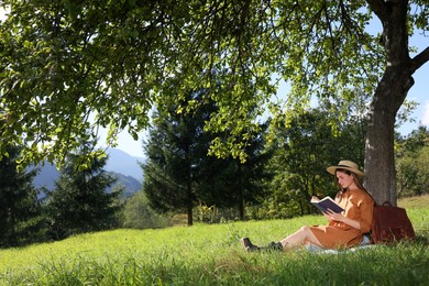 Photo of Young woman reading book under tree on meadow in mountains