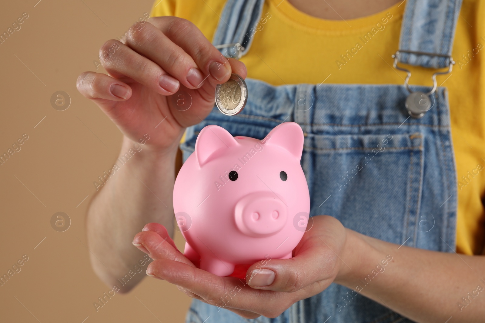 Photo of Woman putting coin into pink piggy bank on light brown background, closeup