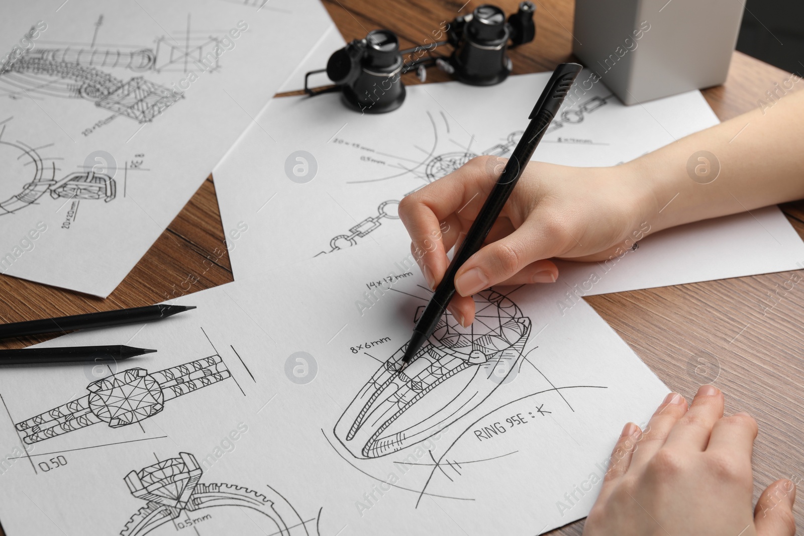 Photo of Jeweler drawing sketch of elegant ring on paper at wooden table, closeup