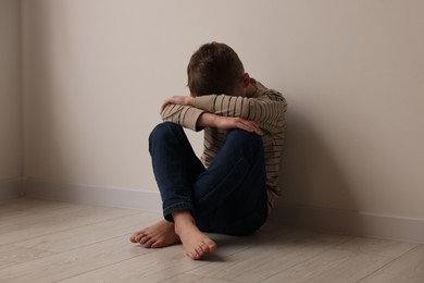 Photo of Child abuse. Upset boy sitting on floor near beige wall indoors