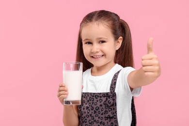 Photo of Cute girl with glass of fresh milk showing thumb up on pink background