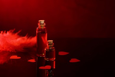 Photo of Bottles of love potion, paper hearts and feather on mirror surface against dark background, space for text. Red tone effect
