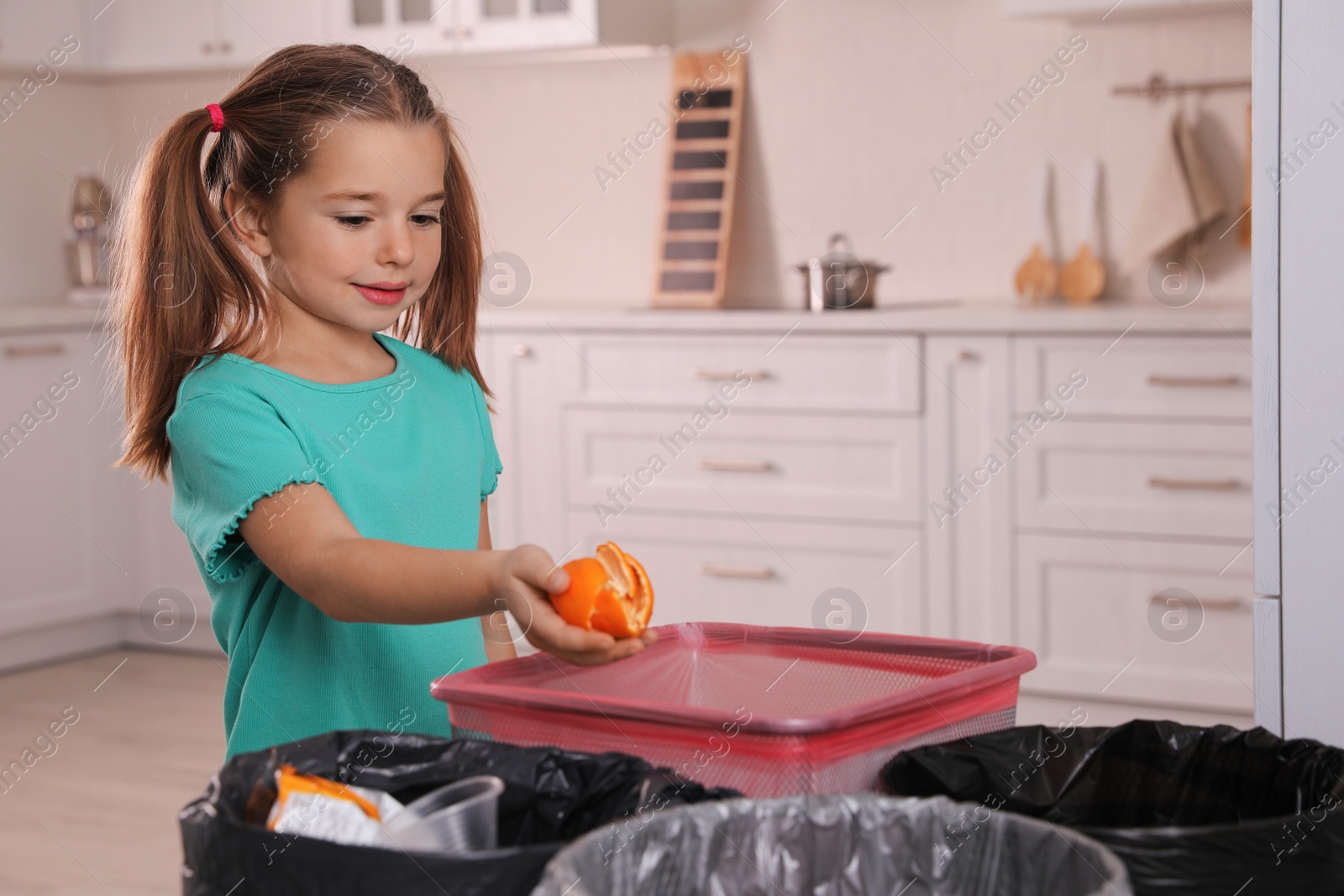 Photo of Little girl throwing tangerine peel into trash bin in kitchen. Separate waste collection