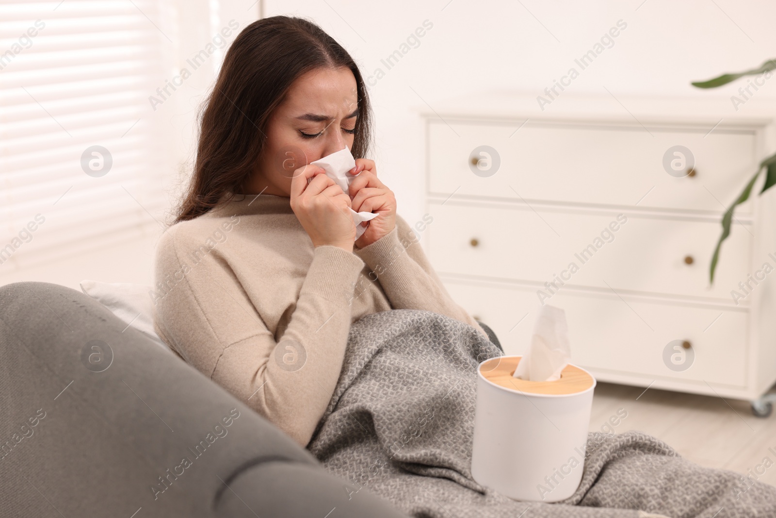 Photo of Sick woman with tissue blowing nose on sofa at home. Cold symptoms
