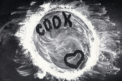 Photo of Heart and word COOK written on flour scattered over black table, top view