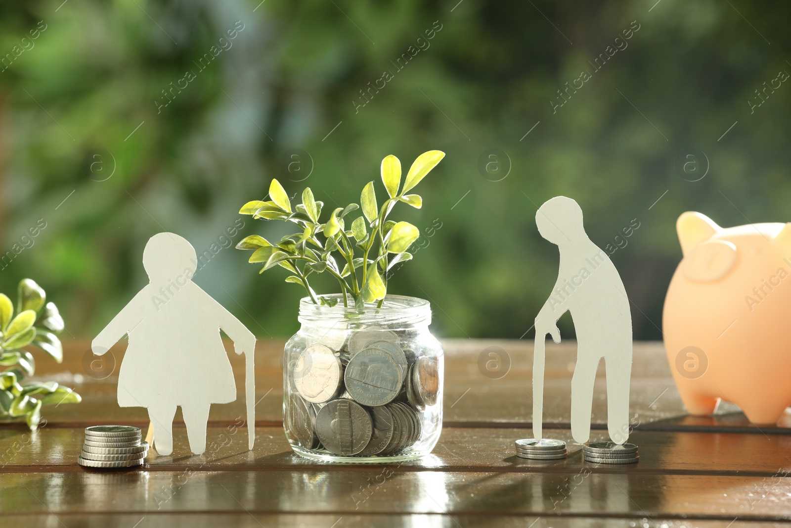 Photo of Pension savings. Figures of senior couple, jar with coins, piggy bank and green twigs on wooden table outdoors