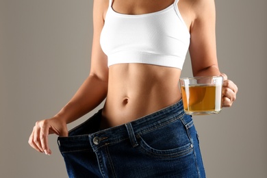 Photo of Young woman in old big jeans with cup of tea showing her diet results on beige background, closeup