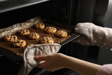 Photo of Woman taking out delicious chocolate chip cookies from oven, closeup