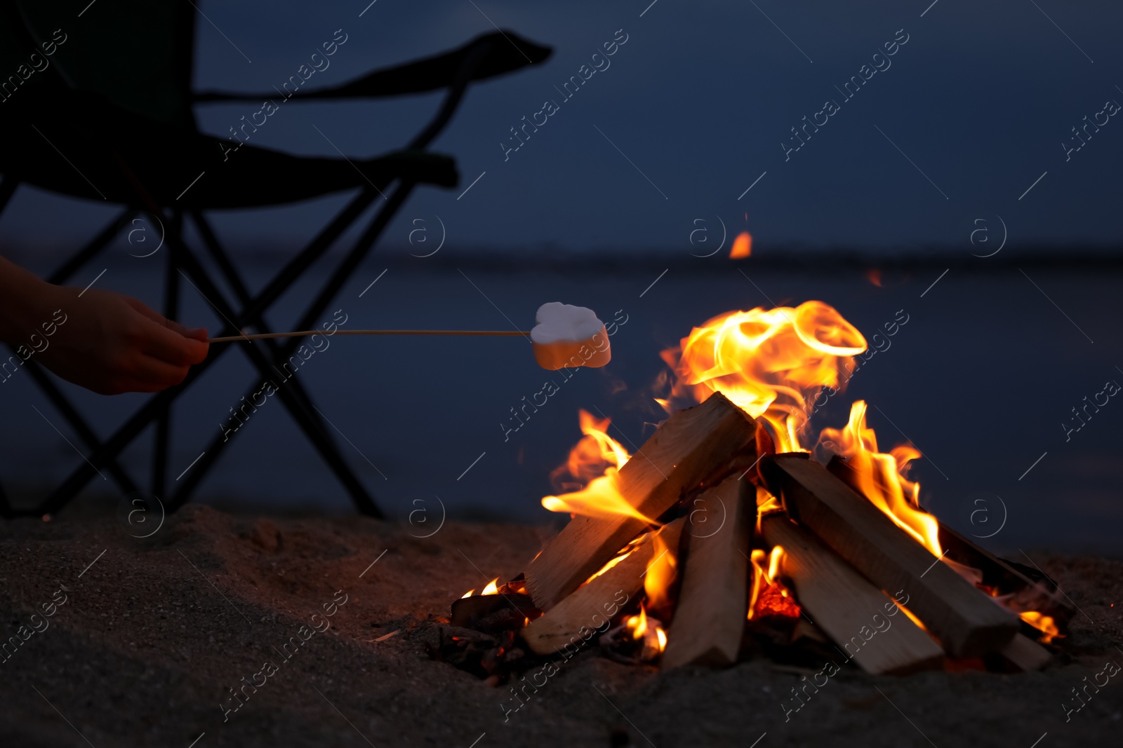 Photo of Woman roasting marshmallow over burning firewood on beach, closeup