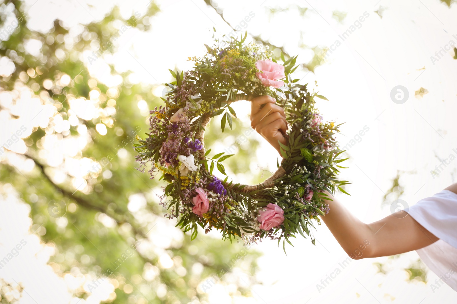 Photo of Young woman holding wreath made of beautiful flowers outdoors on sunny day, closeup