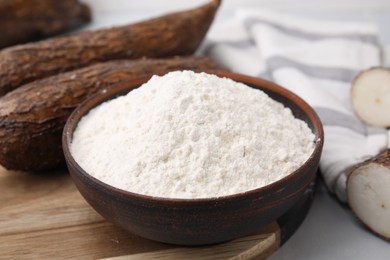 Photo of Bowl with cassava flour and roots on white table, closeup