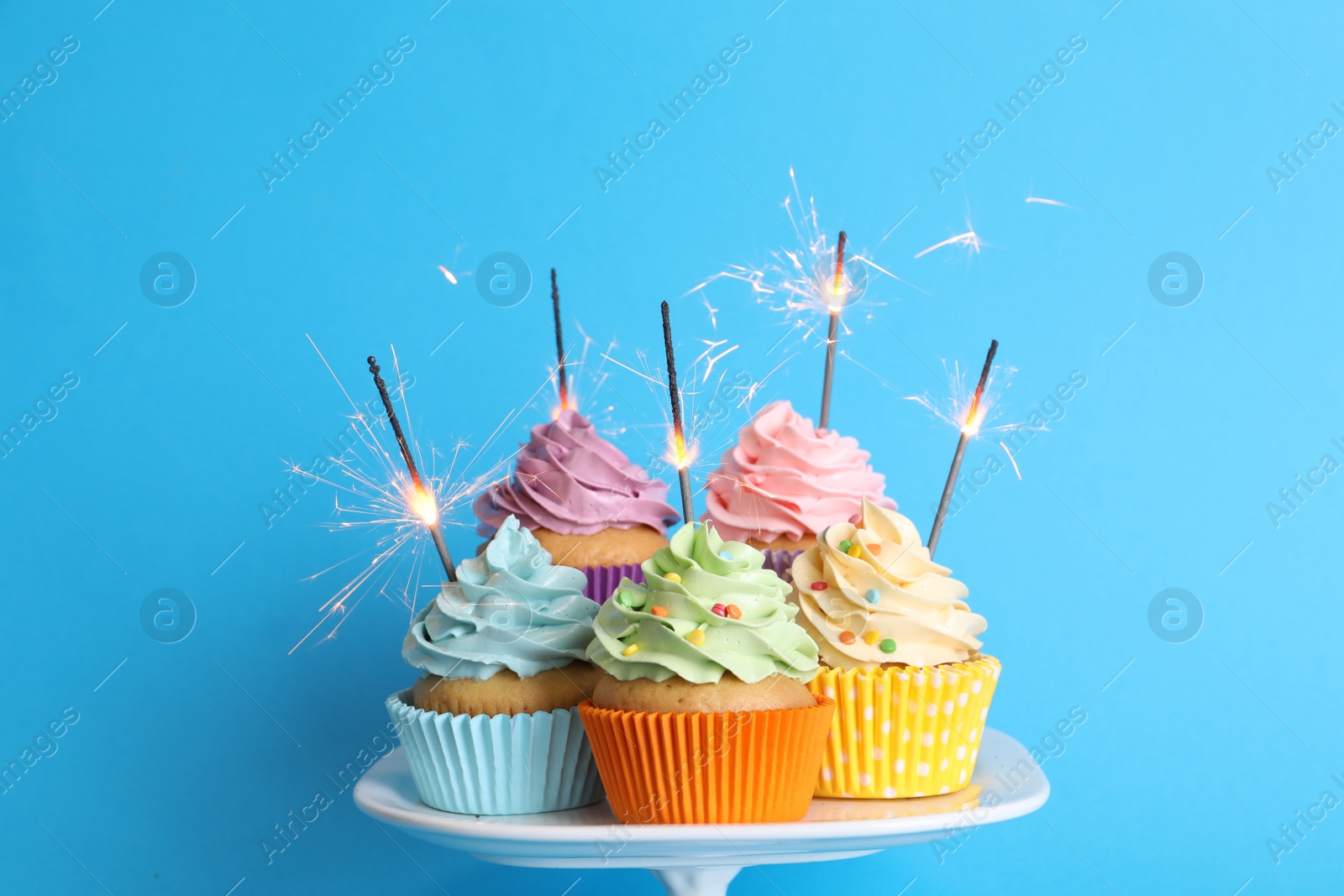 Photo of Birthday cupcakes with burning sparklers on stand against light blue background