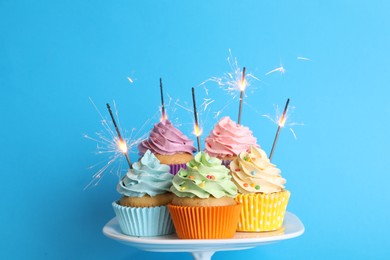 Photo of Birthday cupcakes with burning sparklers on stand against light blue background