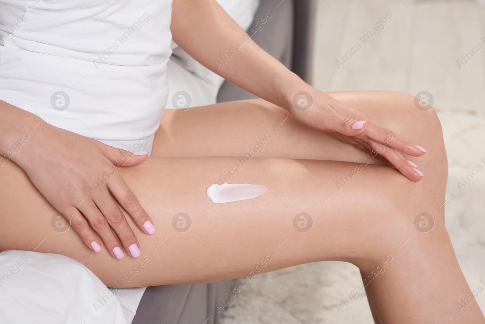 Photo of Woman applying body cream onto her smooth legs in bedroom, closeup