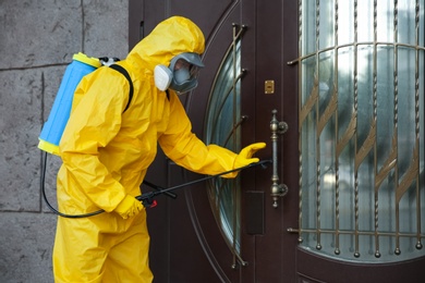 Photo of Person in hazmat suit disinfecting entrance door with sprayer. Surface treatment during coronavirus pandemic