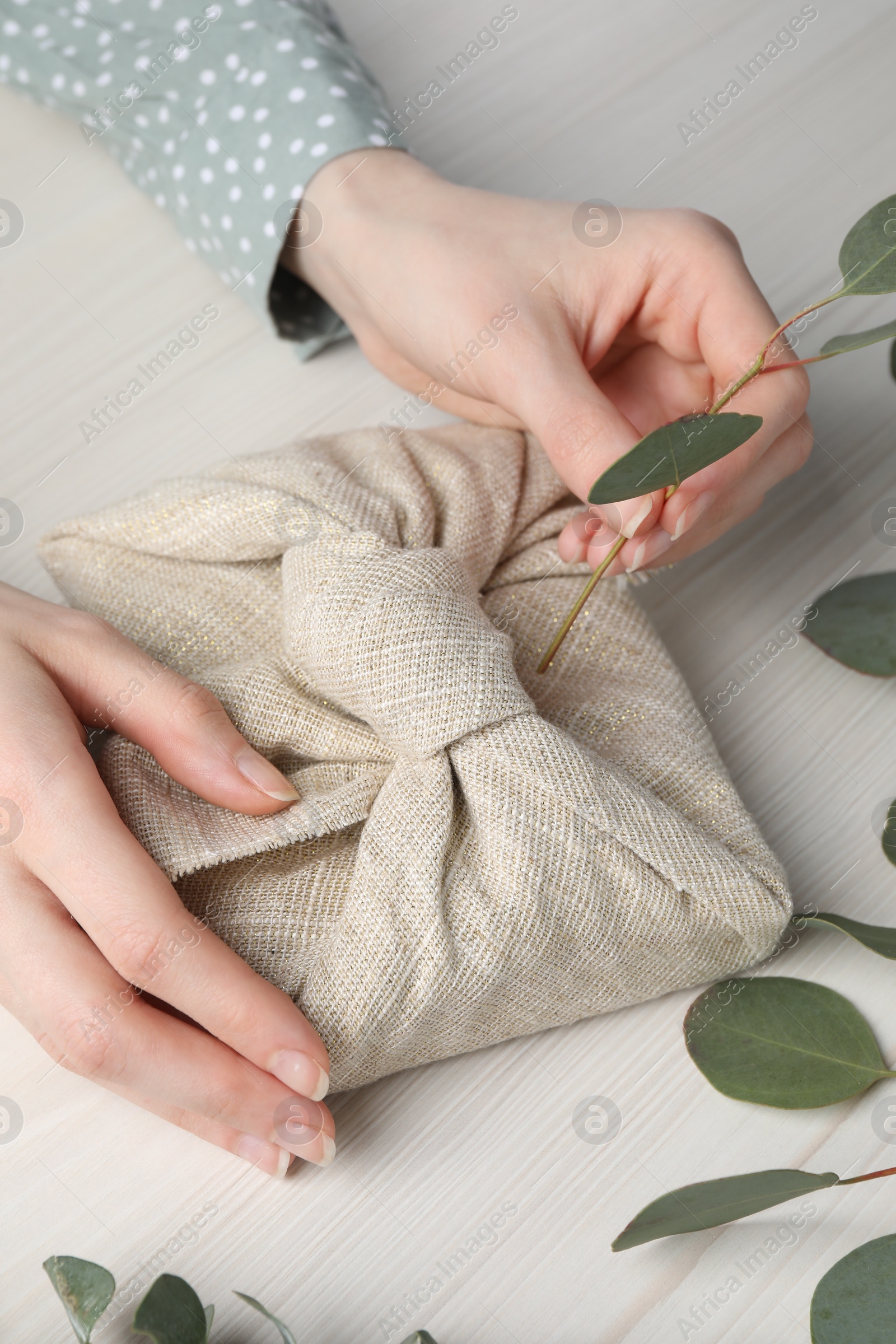 Photo of Furoshiki technique. Woman decorating gift wrapped in fabric with eucalyptus branch at white wooden table, closeup