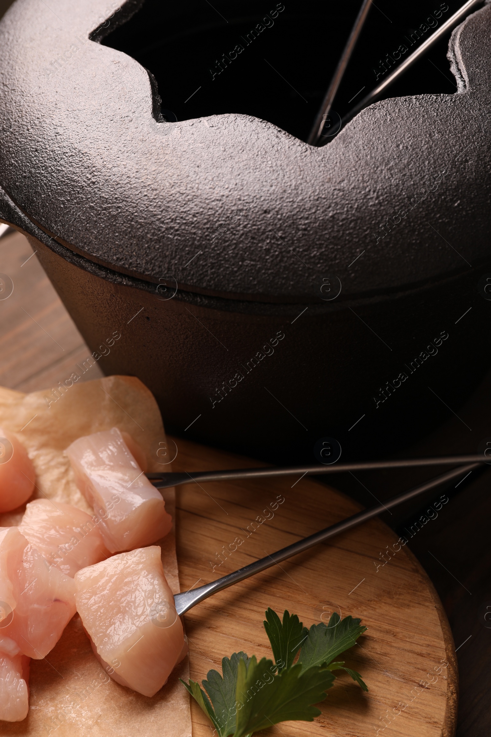 Photo of Fondue pot with oil, forks, raw meat pieces and parsley on wooden table, closeup