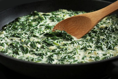 Tasty spinach dip with wooden spoon in frying pan, closeup view