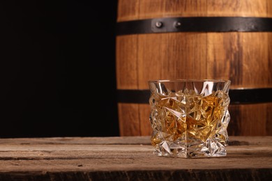 Photo of Whiskey with ice cubes in glass and barrel on wooden table against black background, closeup. Space for text