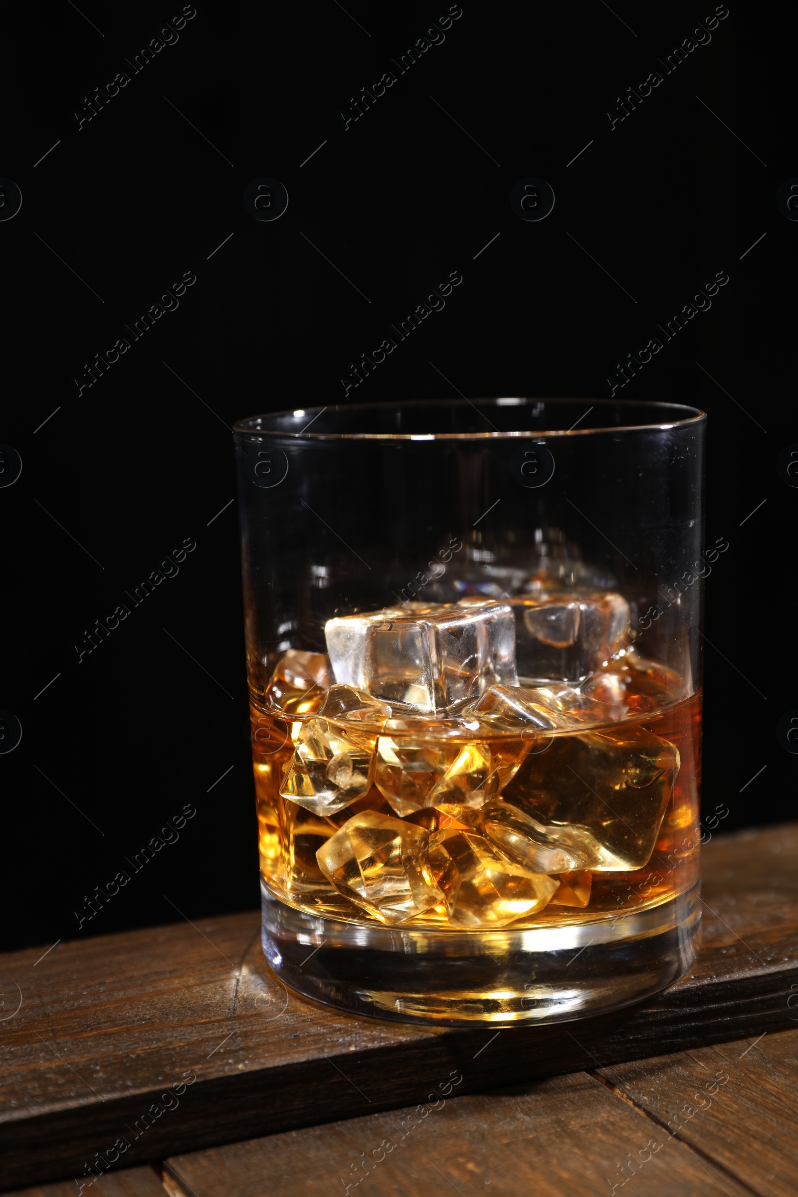Photo of Whiskey with ice cubes in glass on wooden crate against black background, closeup