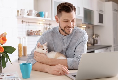 Photo of Young man with cute cat and laptop at home