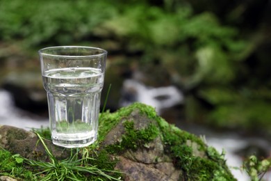 Photo of Glass of fresh water on stone with moss near stream. Space for text