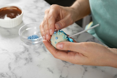 Photo of Young woman decorating tasty cake pop with blue sprinkles at white marble table, closeup