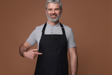 Photo of Happy man pointing at kitchen apron on brown background. Mockup for design