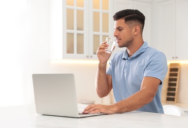 Photo of Man drinking pure water from glass while working with laptop in kitchen