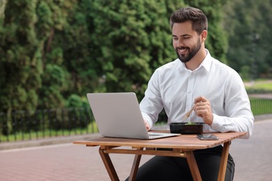Photo of Happy businessman using laptop while having lunch at wooden table outdoors