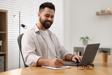 Photo of Young man writing down notes during webinar at table in room