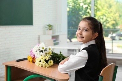 Happy schoolgirl with bouquet sitting at desk in classroom. Teacher's day