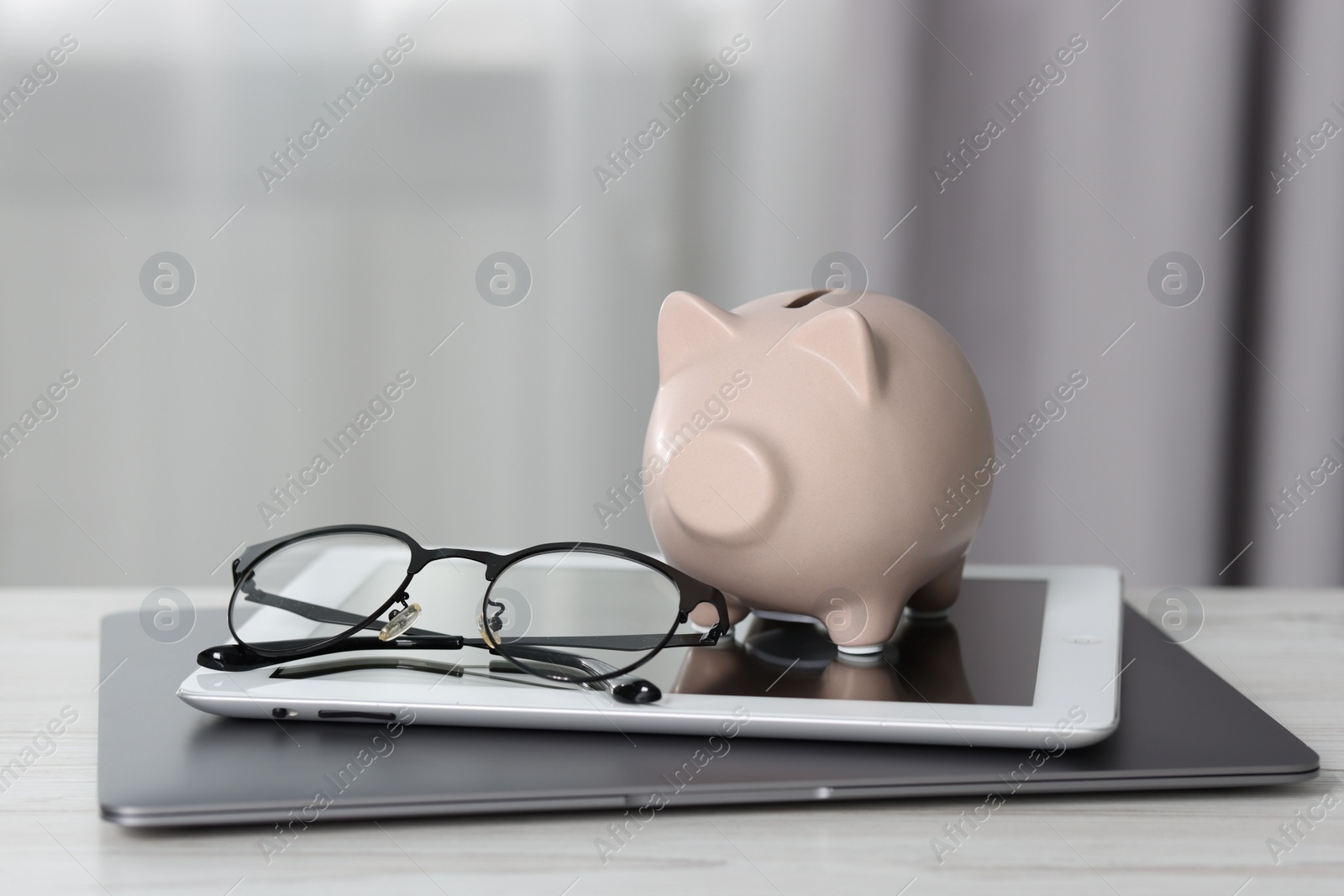 Piggy bank, glasses, tablet and laptop on white table indoors Photo of Piggy bank, glasses, tablet and laptop on white table indoors