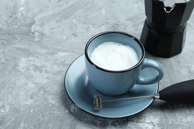Mini mixer (milk frother), whipped milk in cup and moka pot on grey textured table. Space for text