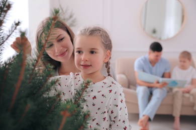 Photo of Mother with daughter decorating Christmas tree together while father and son sitting on sofa at home