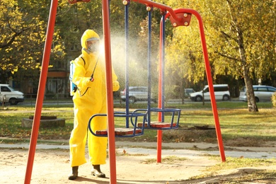 Photo of Person in hazmat suit with disinfectant sprayer cleaning children's playground. Surface treatment during coronavirus pandemic