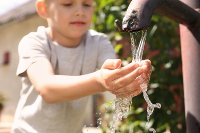 Photo of Water scarcity. Cute little boy drawing water with hands from tap outdoors, selective focus