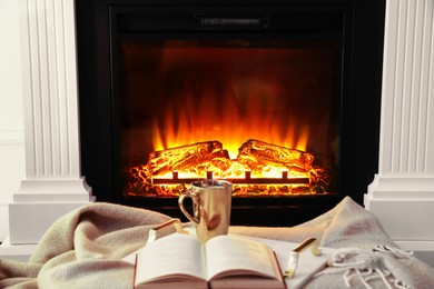 Photo of Cup of hot drink and book on blanket near fireplace indoors. Magic atmosphere