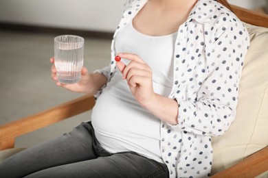 Photo of Pregnant woman holding pill and glass of water indoors, closeup