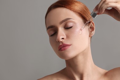Photo of Beautiful young woman applying cosmetic serum onto her face on grey background, space for text