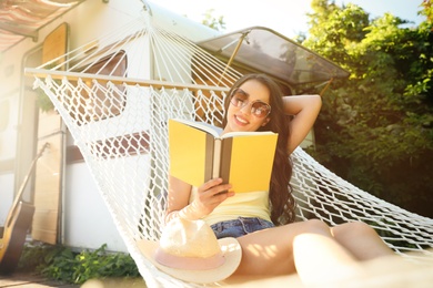 Photo of Young woman reading book in hammock near motorhome outdoors on sunny day
