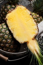 Whole and cut ripe pineapples in metal basket on black table, top view