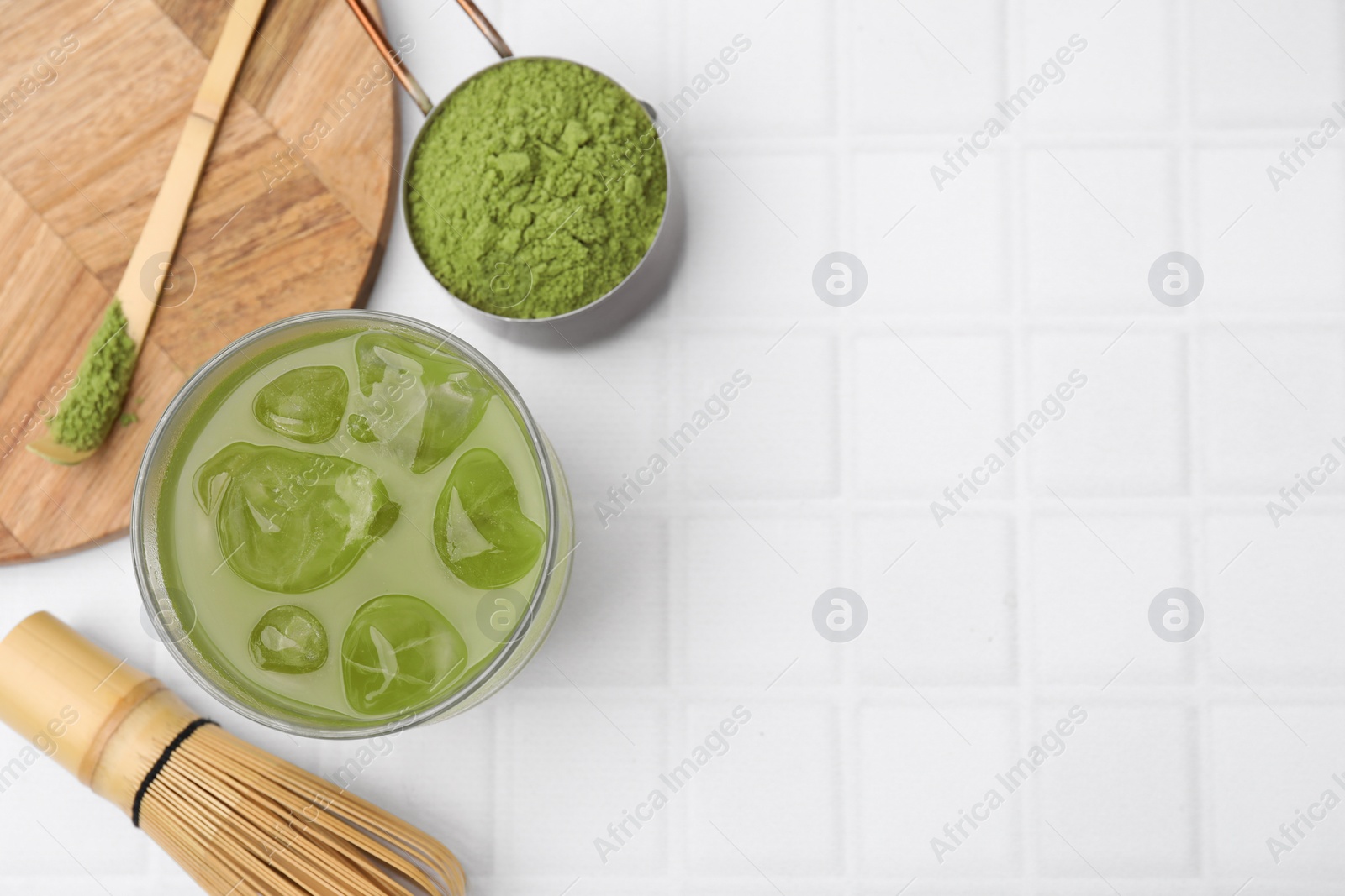 Photo of Glass of delicious iced green matcha tea, powder, bamboo whisk and spoon on white tiled table, flat lay. Space for text