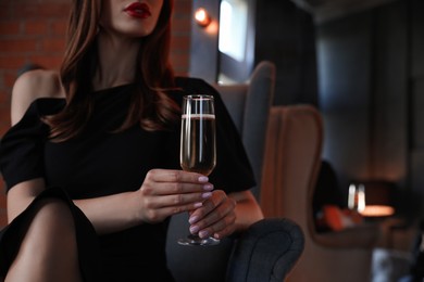 Photo of Elegantly dressed woman with glass of champagne in armchair indoors, closeup. Luxury lifestyle
