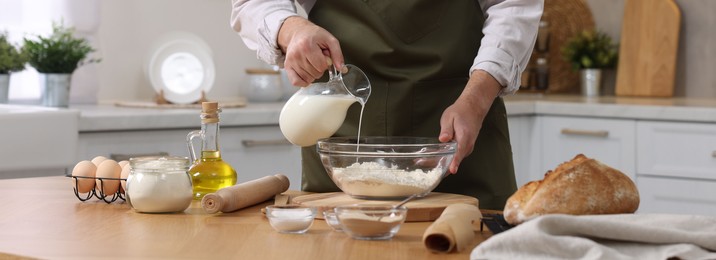 Photo of Making bread. Man pouring milk into bowl with flour at wooden table in kitchen, closeup